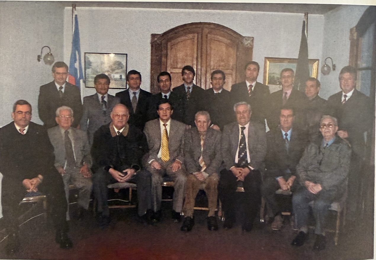 Foto grupal de los alguaciles del círculo en 2004, sentados y de pie frente a una puerta de madera en arco, entre banderas